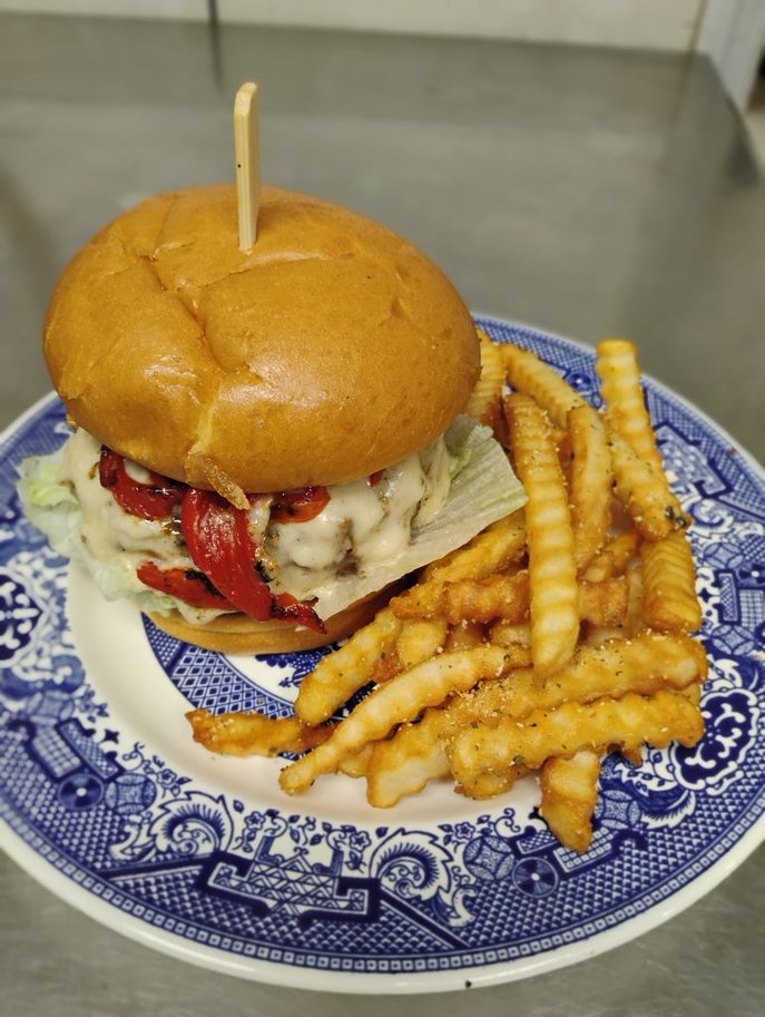 The Red White and Blue, July 2025 Burger of the Month, a burger with melted beer cheese, lettuce, and roasted red peppers on a toasted brioche bun, served with crinkle-cut fries on a blue and white plate